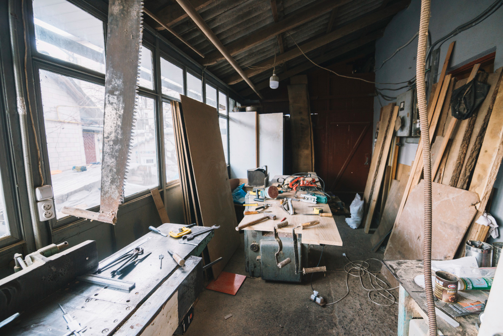A cluttered woodworking workshop with various tools, wooden boards, and equipment scattered on a workbench and floor. Natural light filters in through windows.