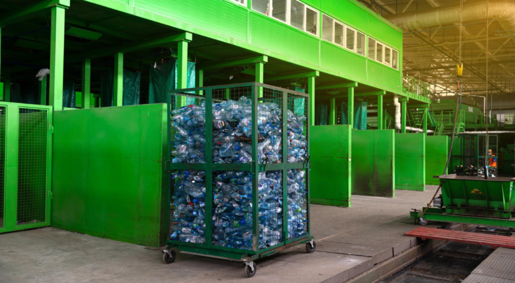 A metal cart filled with plastic bottles stands in a green recycling facility, with green walls and work areas in the background.